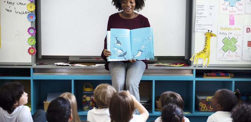 sala de aula com uma professora negra segurando um livro aberto ensinando crianças